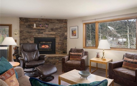 A cozy living room with a stone fireplace, two brown armchairs, a recliner, a sofa, and a large window with a view of the outdoors.