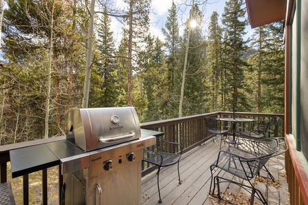 A wooden deck with a gas grill and a dining set, overlooking a forest with tall trees.