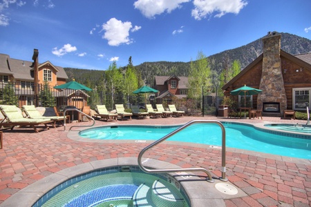 Outdoor pool area with lounge chairs, umbrellas, a mountain view, and a hot tub in the foreground. The scene includes a stone fireplace and multiple wooden buildings in the background.