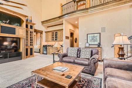 A spacious living room with beige walls, a high ceiling, and wooden furnishings. It features a large couch, a flat-screen TV, a rustic coffee table, and an open kitchen with wooden cabinets.