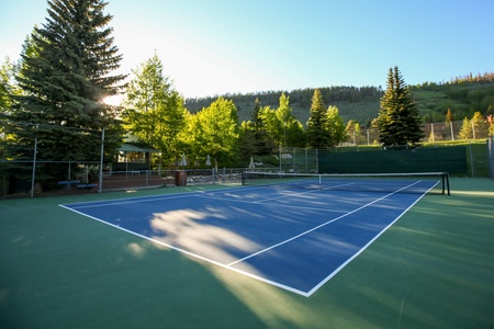 A tennis court surrounded by trees and hills, illuminated by sunlight in the early morning or late afternoon.