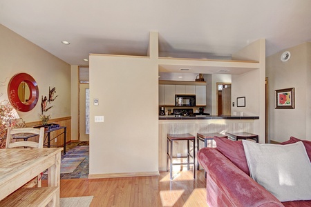 A modern, open-concept living space with a kitchen, dining area, and seating area. The kitchen features a breakfast bar with two stools, and light wood flooring and neutral walls dominate the area.