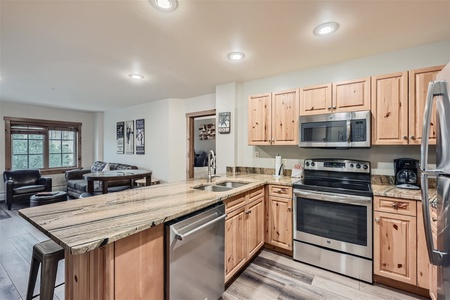 A modern kitchen with wooden cabinets, an island with a granite countertop, stainless steel appliances, and a view into the living area with a dining table and couch.