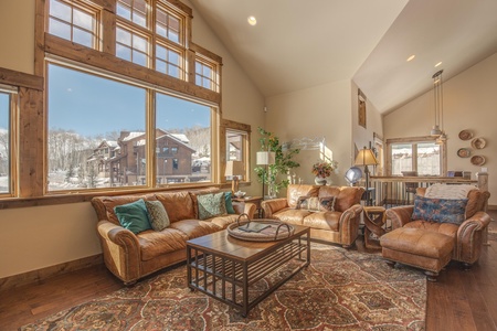 A cozy living room with leather sofas, large windows showing a snowy exterior, wooden furniture, and a patterned rug. The room has high ceilings and is decorated with plants and lamps.