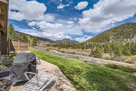 View of a grassy backyard with a barbecue grill and lounge chairs overlooking a scenic landscape featuring mountains, trees, and a partly cloudy sky.