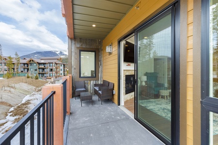 A balcony with two wicker chairs overlooks a scenic view of a residential area with mountains in the background. The balcony is attached to a yellow building with a sliding glass door leading inside.