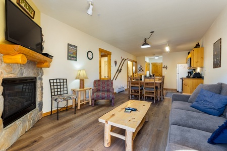 A cozy living room with a fireplace, TV, sofa, chairs, wooden coffee table, and dining area. Kitchen with appliances is visible in the background. Decor includes wall art, a clock, and skis.