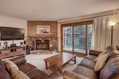 A living room with a stone fireplace, large sectional leather sofa, wooden coffee table, wall-mounted TV, and sliding glass doors leading to an outdoor balcony with a forest view.