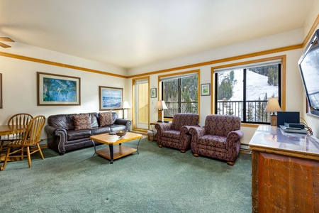 A cozy living room with green carpet, brown leather sofa, two patterned armchairs, and a wooden dining table with chairs. Large windows reveal a snowy landscape outside. Wall art decorates the space.