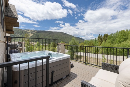 A deck with a hot tub, outdoor seating, and a railing overlooks a forested mountain landscape under a partly cloudy sky.