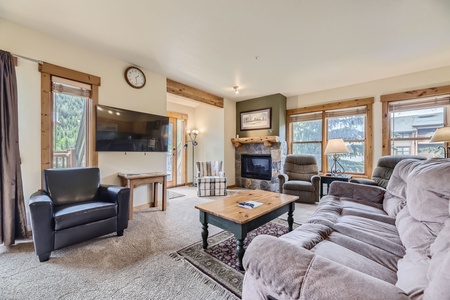 A cozy living room with a sofa, armchairs, a coffee table, a fireplace, a wall-mounted TV, and large windows offering a mountain view. There is a clock on the wall and a small rug on the floor.