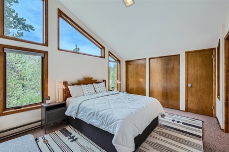 A bedroom with angled windows, wooden accents, and a white bedspread. The room features a minimalist design with two patterned rugs and natural light streaming in through the windows.