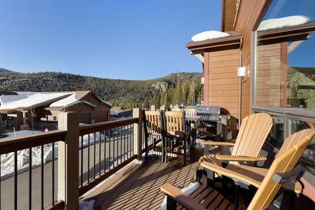 Outdoor deck with wooden chairs and a barbecue grill overlooking a snowy mountain and forest landscape. Cabin-style buildings are visible in the background under a clear blue sky.