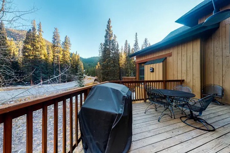 Wooden deck with a grill, table, and chairs, overlooking a scenic view of tall pine trees and mountains.