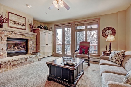 A cozy living room features a stone fireplace, wooden coffee table, sofa, armchair, ceiling fan, and large windows with a view of trees. A lamp and a wall-mounted clock adorn the space.