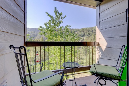 A small balcony with two green-cushioned chairs, a round metal table, and a view of a lush green hillside and trees.