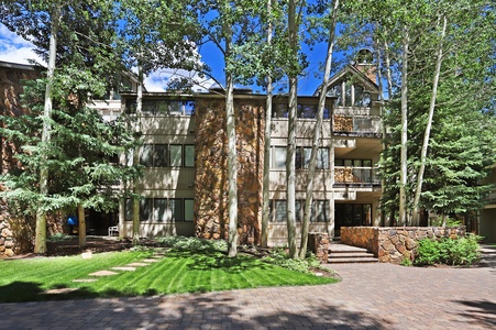 A multi-story building with stone and wood exterior, surrounded by trees and featuring a paved walkway and green lawn in front.