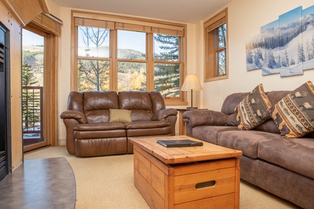 A cozy living room with two leather couches, a wooden coffee table, a floor lamp, and wall art depicting snowy landscapes. Large windows offer a view of trees and hills outside.