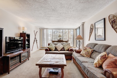 A living room with a rustic theme featuring a brown sofa, a wooden coffee table, two armchairs, a flat-screen TV on a stand, and wall-mounted skis and snowshoes. Large windows provide natural light.