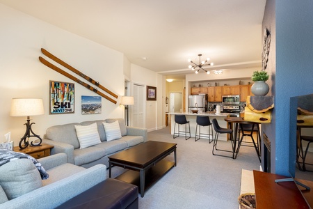 A modern living room with a gray sofa, wooden coffee table, wall-mounted skis, and a kitchen area in the background featuring light wood cabinets and stainless steel appliances.