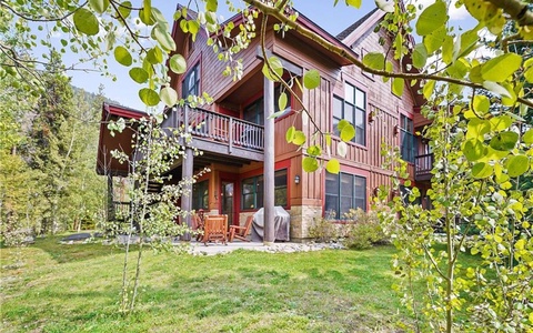 A two-story wooden house with balconies and large windows is surrounded by green trees and grass in a scenic outdoor setting.