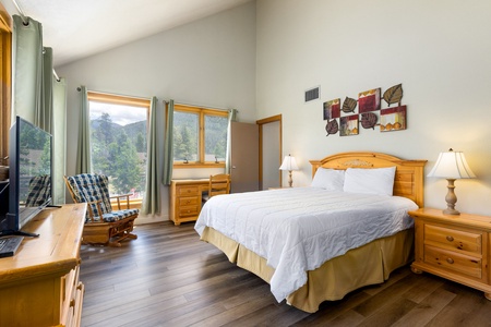 A bedroom with a wooden bed, nightstands, a TV, an armchair, and large windows with green curtains. The walls are decorated with leaf-themed art. The floor is hardwood, and there is a mountain view outside.