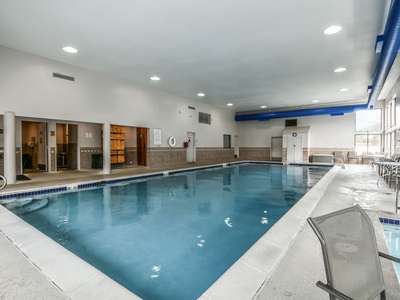 An indoor swimming pool with blue water, surrounded by white and beige tiles. There are poolside chairs, windows on one side, and doors and wall-mounted equipment on the opposite side.