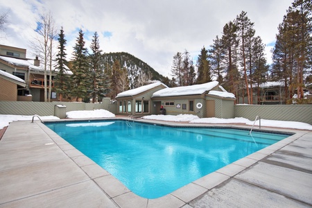 Outdoor swimming pool surrounded by snowy landscape with trees and buildings in the background.