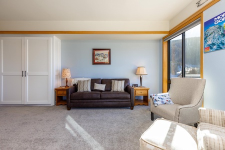 A living room with a brown sofa, two wooden side tables with lamps, an armchair, and a large window. The walls are light blue with a framed painting and a poster.