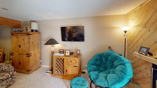 A living room with a TV mounted on the wall, wooden furniture, a teal upholstered round chair with an ottoman, a standing lamp, and family photos on a side table.