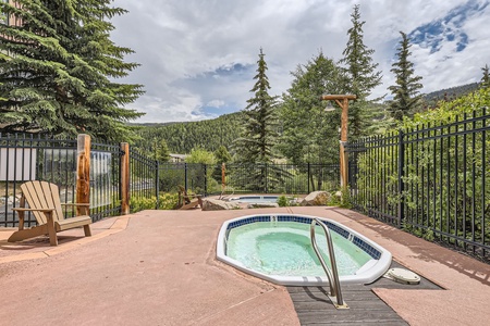 A small outdoor hot tub with a metal handrail, surrounded by a black metal fence, two wooden chairs, and lush green trees against a mountainous backdrop under a partly cloudy sky.