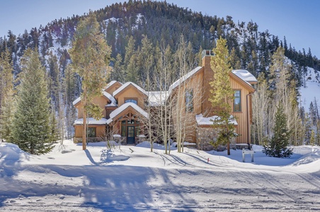 A wooden house with snow-covered roof and trees stands in front of a forested hill in winter.