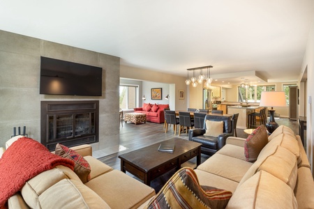 A spacious living room with a beige sectional sofa, armchairs, a coffee table, and a wall-mounted TV above a fireplace. The open-plan space extends to a dining area and a modern kitchen in the background.