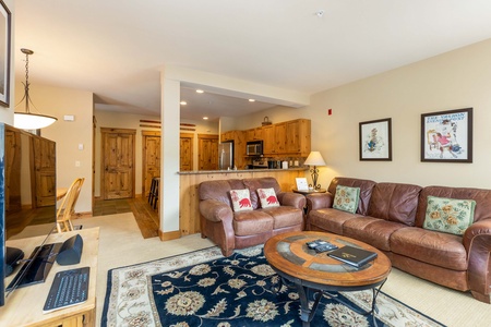 Modern living room with brown leather couches, wooden coffee table, rug, and open view of a kitchen with wooden cabinets and a breakfast bar. Decor includes framed pictures and various pillows.