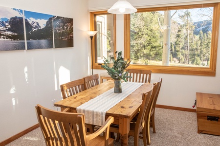 A dining room with a wooden table and chairs, a table runner and vase with flowers. A floor lamp stands in the corner, and large windows offer a view of trees and a mountain landscape. A triptych hangs on the wall.