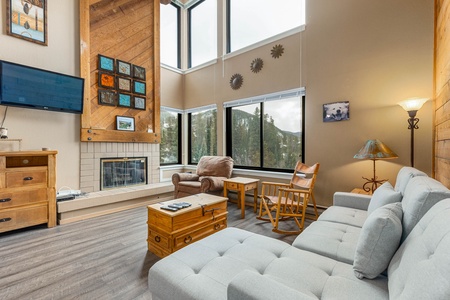 A cozy living room with large windows, a gray couch, a rocking chair, a wall-mounted TV, and a fireplace. The room is decorated with artwork and cushioned furniture.