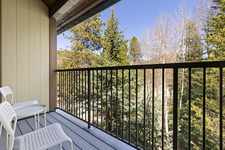 A balcony with two white chairs overlooks a forested area with evergreen trees under a clear sky.