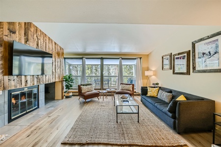 A bright living room with a grey sofa, glass coffee table, two chairs, a fireplace, a large wall-mounted TV, and floor-to-ceiling windows with curtains leading to a balcony.