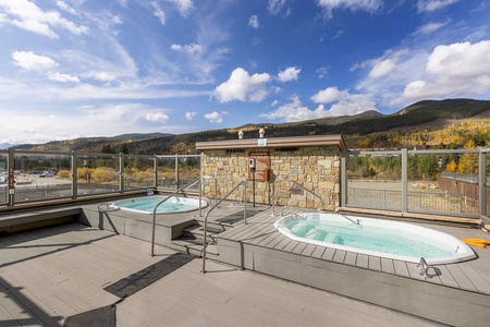 Two outdoor hot tubs on a wooden deck with mountain views in the background. The area is surrounded by clear glass railings and there is a stone wall in the center. The sky is partly cloudy.