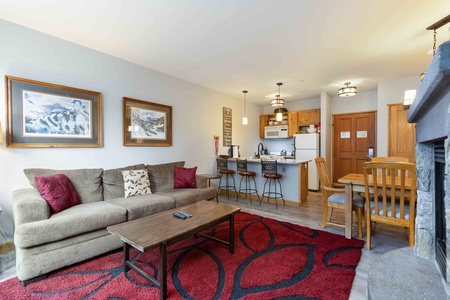 A cozy living room with a brown sofa, coffee table, red and black rug, and kitchen area with counter seating. A wooden dining table is on the right, and two framed paintings are on the wall.