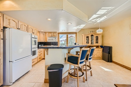 A kitchen with light wood cabinetry, a white refrigerator, an oven, a central island with a dark countertop, and three bar stools. A china cabinet and a window providing natural light are in the background.