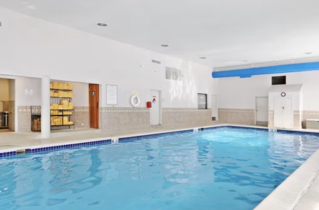 Indoor swimming pool area with tiled walls, a stack of yellow towels on shelves to the left, a lifebuoy hanging on the wall, and blue ductwork on the ceiling.