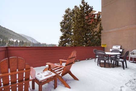 A snowy outdoor patio with wooden chairs, a table with plastic chairs, and a barbecue grill. Pine trees and a mountainous landscape are in the background.