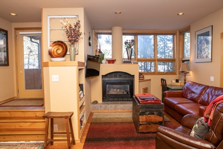 A cozy living room with a fireplace, a brown leather sofa, a red throw blanket, a wooden chest coffee table, and a small desk by the window. Shelves hold decorative items and there is a view outside.