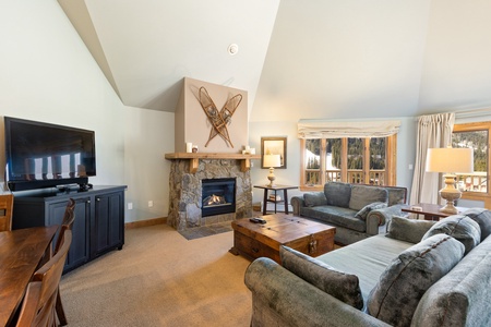 A cozy living room with a stone fireplace, TV, two green sofas, a wooden coffee table, and a scenic view through large windows. Skis are mounted decoratively above the fireplace.