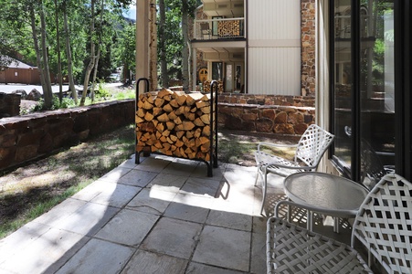 A stone patio with white metal chairs and a glass table, next to a firewood storage rack in front of a stone building.