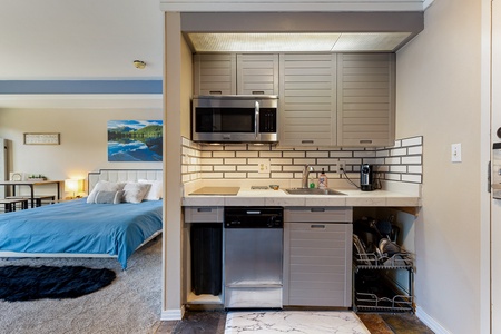 Compact kitchen with stainless steel appliances, a microwave, and a dishwasher, situated next to a bedroom with a blue bedspread and a nature-themed wall art.