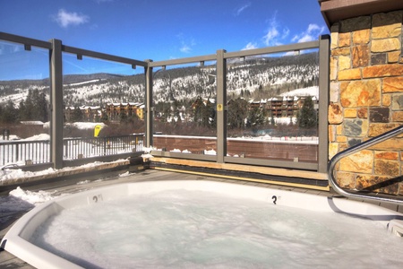 An outdoor hot tub overlooks a snowy landscape with mountains and buildings in the background. The area is enclosed by a glass and metal fence.