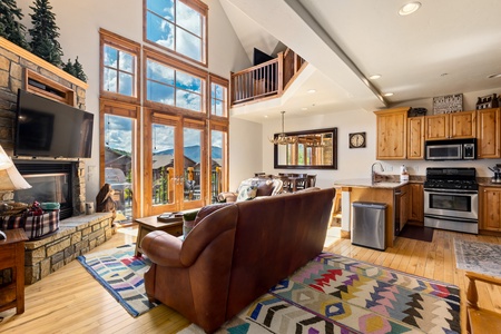 Cozy living room and kitchen with large windows, leather sofas, a stone fireplace, wooden cabinets, and vibrant rugs.