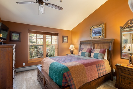 A bedroom with a wooden bed frame, colorful patchwork quilt, dresser, nightstand, table lamp, and an overhead ceiling fan. The walls are painted orange, and a window provides natural light.